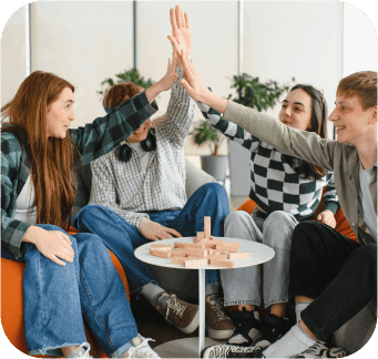 Four cheerful students are giving a high five while sitting on beanbag chairs and playing a board game, demonstrating teamwork and joyful celebration