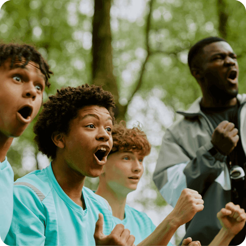 Group of multiethnic teenage boys cheering with excited expressions outdoors, Black man coach standing behind holding digital tablet and shouting encouragement during sports event