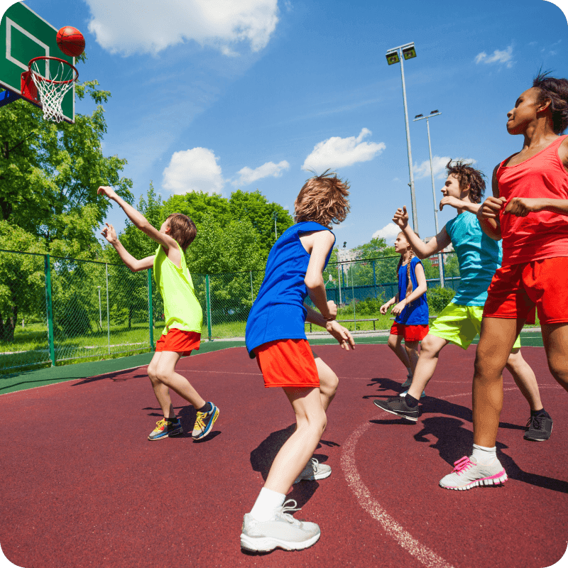Teenagers in colorful uniforms playing basketball game on the ground during sunny summer day together