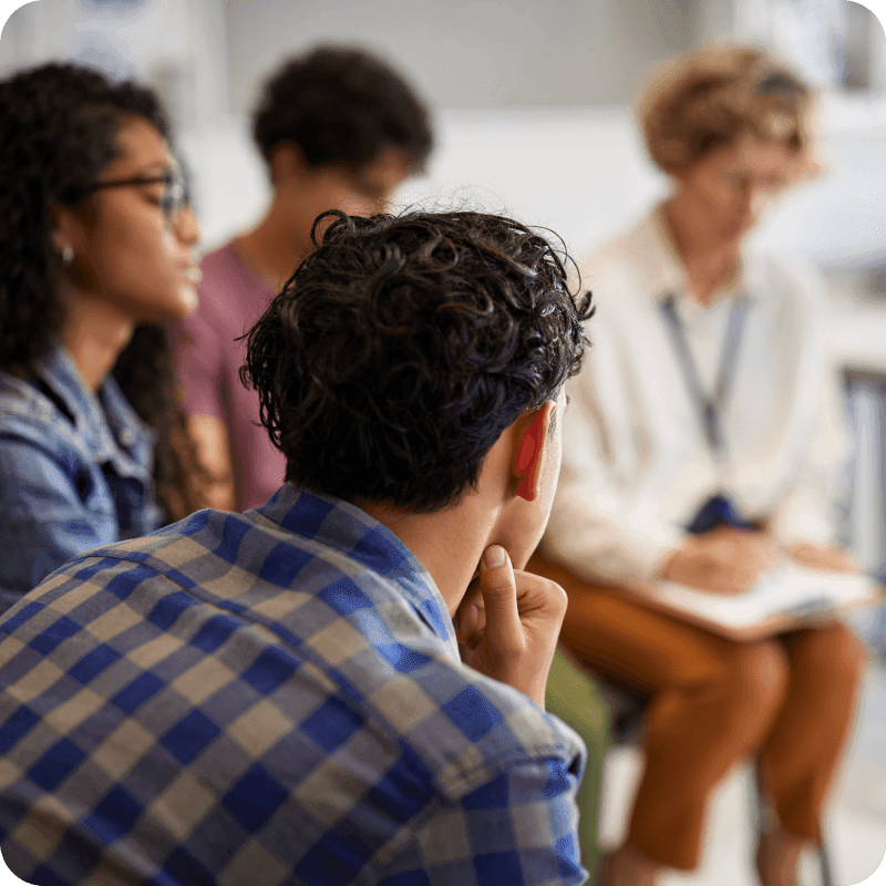 Teenagers sitting in a circle with counselor during a group discussion on emotional well-being. Therapist taking notes while high school students reflect on their feelings and mental health challenges. Multiethnic group of guys and girls listening to therapist during support session.