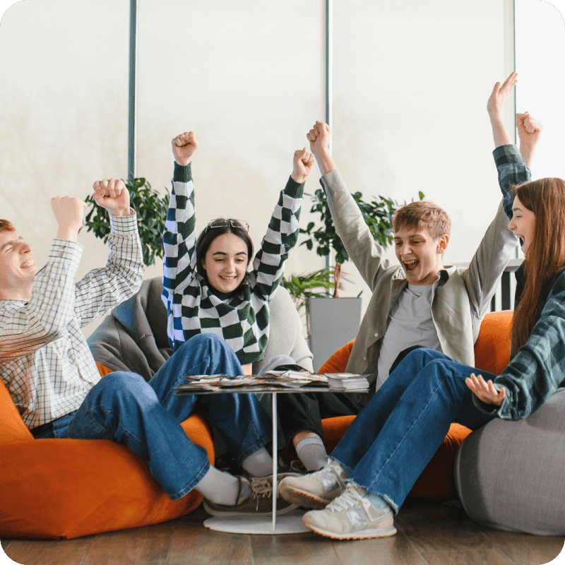Four cheerful students are raising their arms in excitement while playing a board game, enjoying their leisure time together in a modern school area