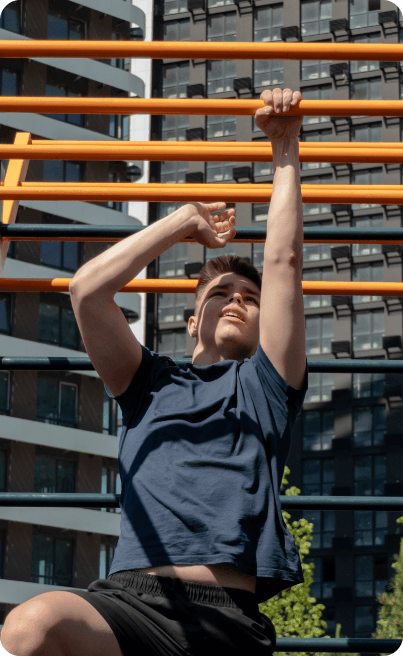 A determined young man engages in an intense physical workout on a set of bright orange monkey bars at a modern outdoor street workout park. His athletic build and focused expression highlight dedication to strength and fitness.